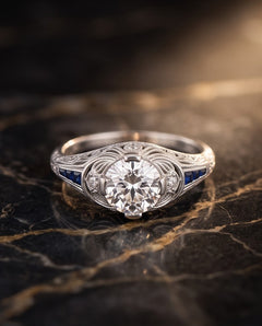 Art Deco style platinum engagement ring with .95 carat round brilliant diamond, French-cut sapphire shoulders and filigree gallery, shown on dark marble